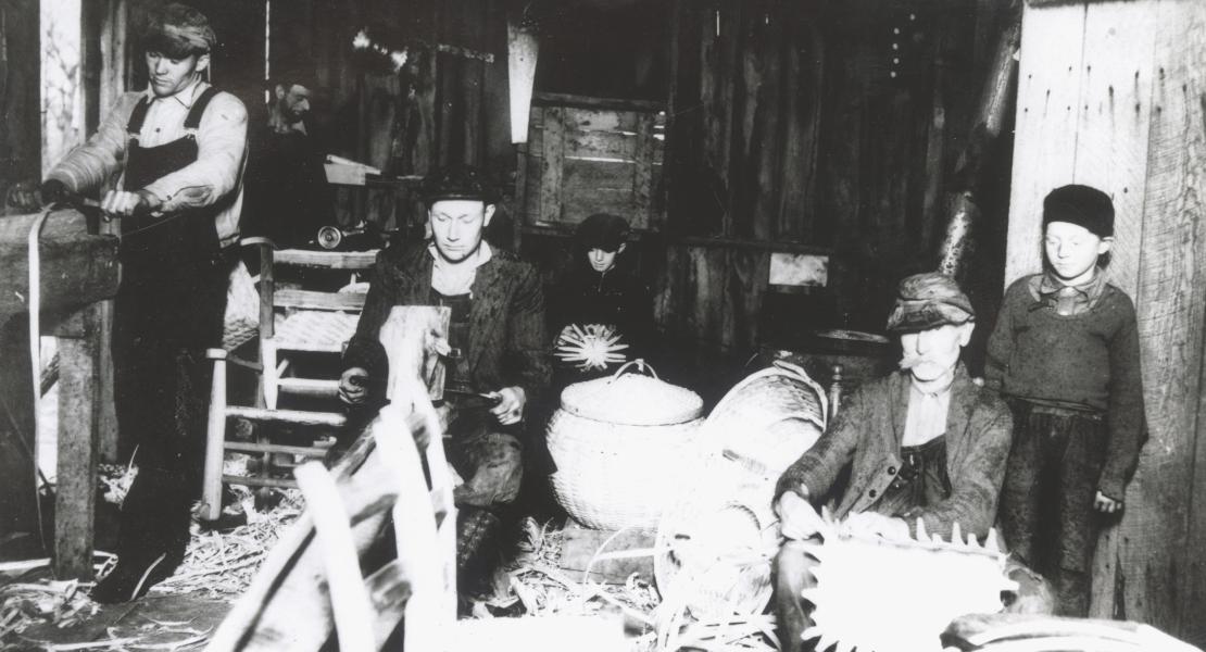 The home-based basket factory run by members of the Childers family (left to right): Harry Thompson, husband of Amy Childers; George Ruckman, husband of Janettie Childers, making chair rungs; Andrew Childers on shaving horse; Chester Ruckman in the background; William F. Childers making basket; George Ruckman Jr. standing by door. [Phelps County Historical Society, Elbert I Childers Collection]